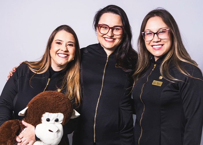 Três integrantes da equipe da Clínica IEM sorrindo, de uniforme preto, uma delas segurando o macaco de pelúcia da odontopediatria
