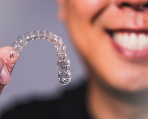 Mão segurando um alinhador transparente, com paciente sorrindo ao fundo