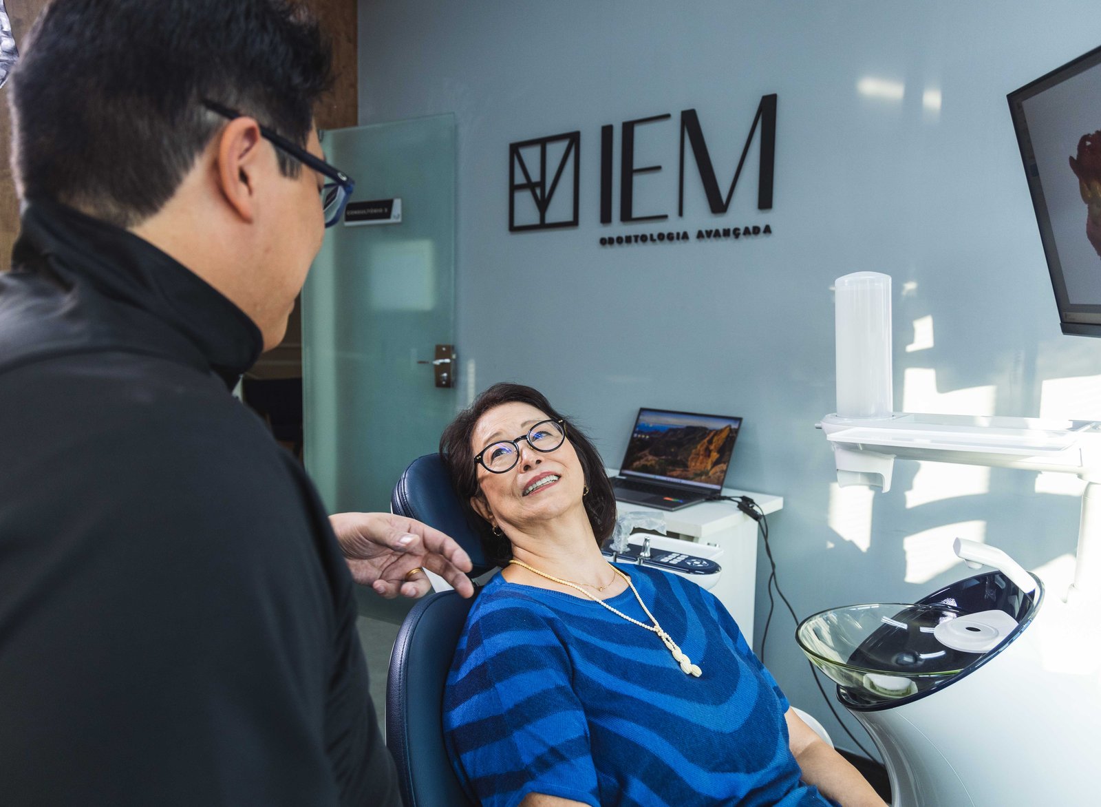 Dr. Willian Akinori Saito conversando com paciente sorridente na cadeira odontológica, sob o logotipo da IEM Odontologia Avançada